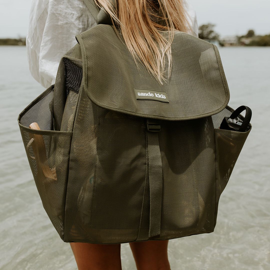 Person wearing a green backpack with 'sande kids' branding by a body of water.