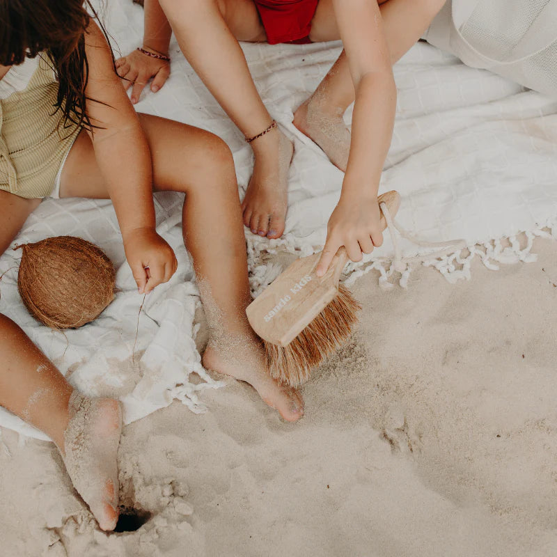 two kids using Sande Kids sand brush to remove sand on beach from legs and feet.