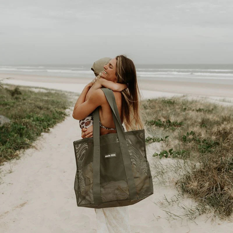 Mum carries Mesh beach bag on shoulder while hugging her toddler at the beach entrance.