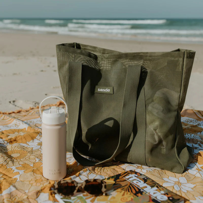 Sande Kids Beach bag in pandanus green sitting on picnic rug on sand at beach.