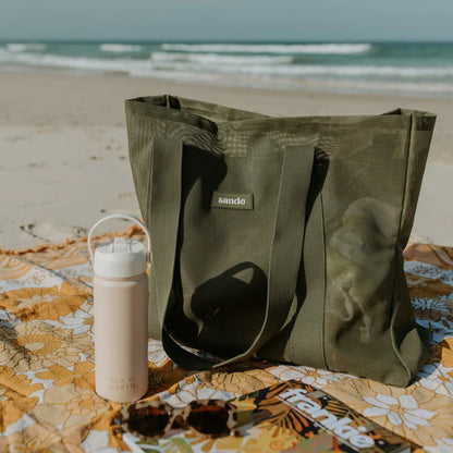 Sande Kids Beach bag in pandanus green sitting on picnic rug on sand at beach.