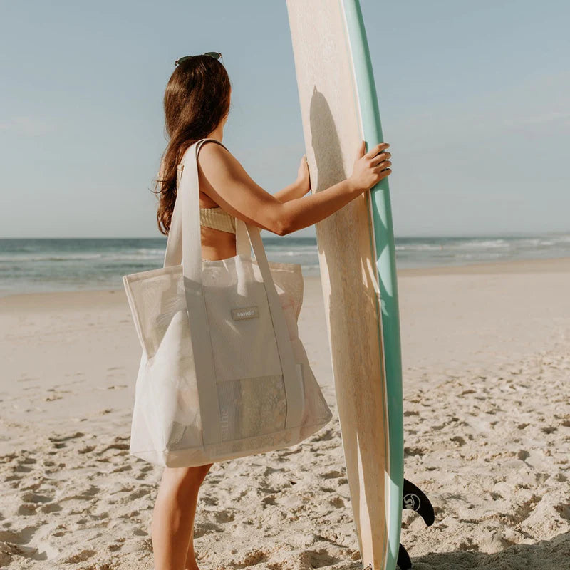 Girl with beach bag on shoulder with surf board on beach.